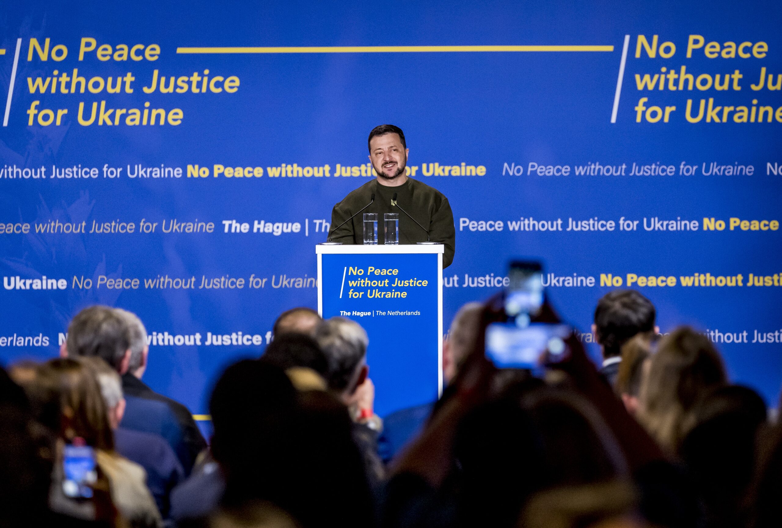 Spreker in militair groen shirt geeft presentatie achter podium met blauwe achtergrond met tekst "No Peace without Justice...